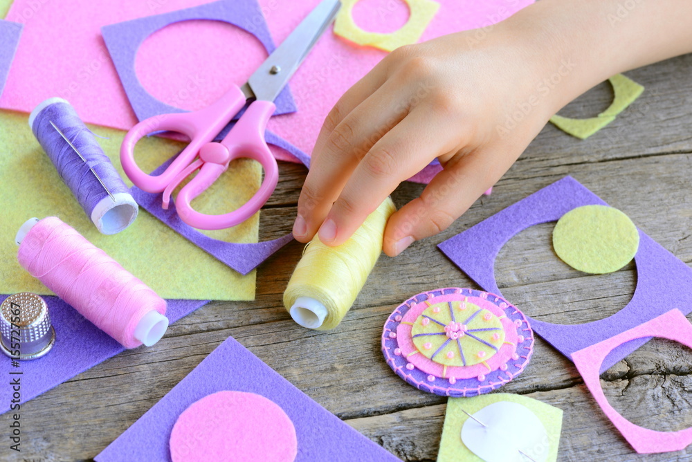 Fototapeta premium Teaching children to sew by hand concept. Small child made a flower from felt circles. Child takes thread in hand. Beaded felt flower crafts, craft supplies on wooden table. Easy and quick applique