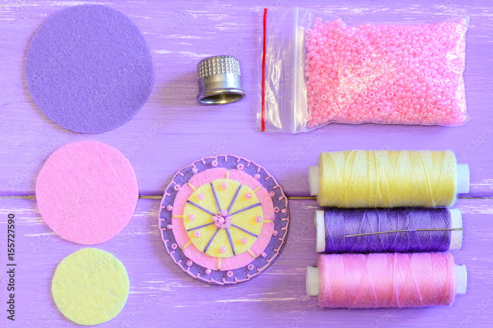 Fototapeta premium Felt flower, colored felt circles, thread spools, needle, thimble, pink beads on a wooden table. Sewing felt flower tutorial. Simple round felt flower crafts for kids. Top view