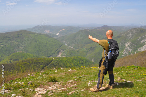 Hiker with backpack relies on a stick and pointing to the direction of the valley. Man on top of the mountain look at the valley below
