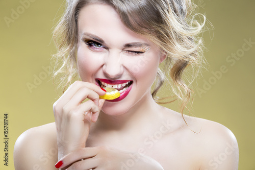 Portrait of Sexy Caucasian Blond Girl Eating Tiny Lemon Piece. Demonstrating Too Much of Acid Taste Of the Fruit. Against Yellow Background.