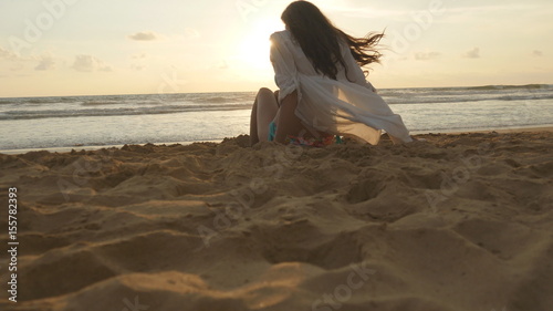 Beautiful young woman sitting on golden sand on sea beach during sunset and calls to himself. Girl relaxing on ocean shore during summer vacation travel at sundown. Ocean waves at background. Close up photo