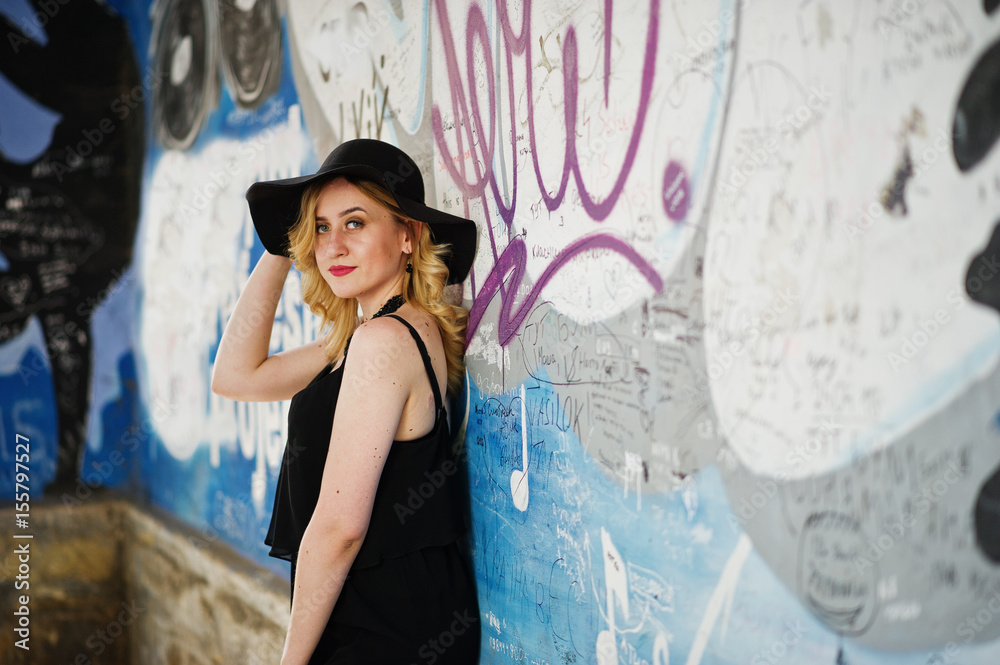 Naklejka premium Blonde woman on black dress, necklaces and hat against graffiti wall.