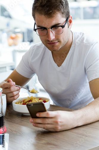Handsome young man using his mobile phone while enjoying the breakfast in the kitchen.