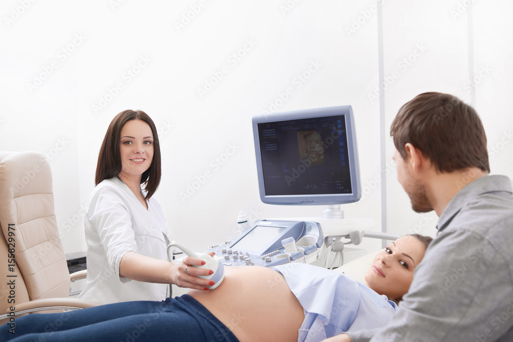 Smiling professional female doctor in white uniform using ultrasound equipment, screening pregnant woman. Nurse touching stomach of woman by tool. Parents expecting baby and diagnosing in clinic. Smiling professional female doctor in white uniform using ultrasound equipment, screening pregnant woman. Nurse touching stomach of woman by tool. Parents expecting baby and diagnosing in clinic.