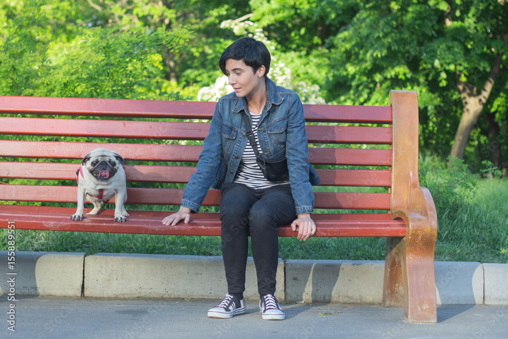 Fototapeta premium Portrait picture of young pretty woman in the park sit on bench, walk of female and pug in the summer park. 