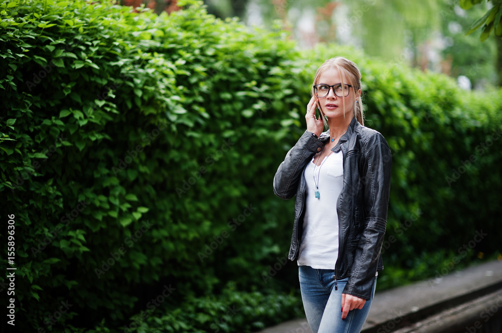 Stylish blonde woman wear at jeans, glasses and leather jacket with phone at hand, against bushes at street. Fashion urban model portrait. Stylish blonde woman wear at jeans, glasses and leather jacket with phone at hand, against bushes at street. Fashion urban model portrait.