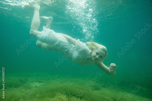 A floating woman. Underwater portrait. Girl in white dress swimming in the lake. Green marine plants and water