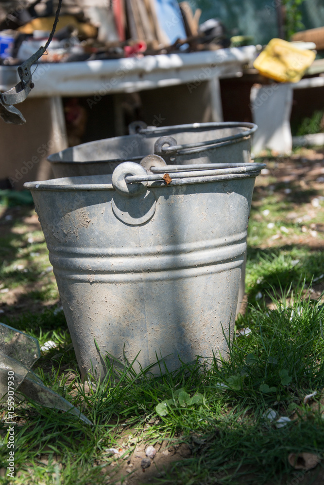 Close up of two rusty iron buckets in grass in the garden. Dirty gray metallic bucket with garbage on barnyard at sunny day. Gardening background. Containers for storing feed, trash, water, paint Close up of two rusty iron buckets in grass in the garden. Dirty gray metallic bucket with garbage on barnyard at sunny day. Gardening background. Containers for storing feed, trash, water, paint
