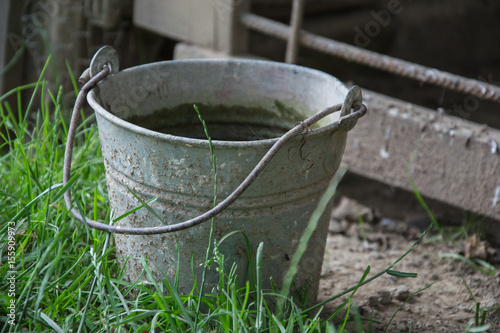 Close up of rusty iron bucket in grass in the garden. Dirty gray metallic bucket with garbage on barnyard at sunny day. Gardening background. Containers for storing feed, trash, water, paint