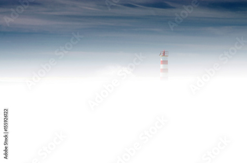 Sylwetka latarni morskiej wyłaniająca się spoza gęstej mgły. Granatowe niebo, gęsta mleczna mgła, drobna sylwetka latarni po prawej stronie. 