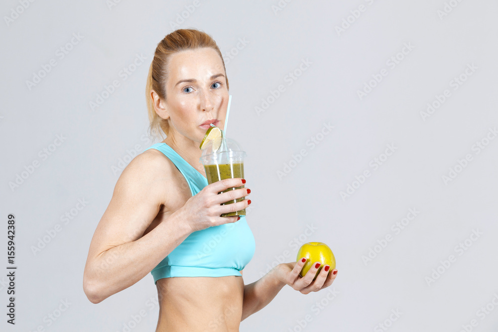 middle-aged athletic slim woman drinking the smoothie and holding an apple on the grey background middle-aged athletic slim woman drinking the smoothie and holding an apple on the grey background