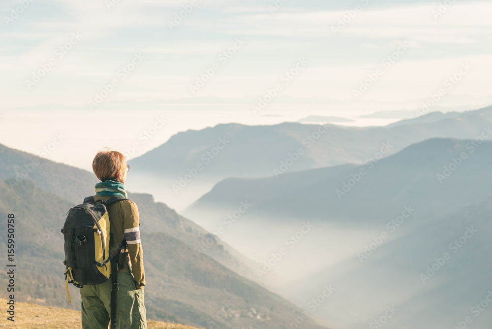 Female hiker with backpack looking at the majestic view on the italian Alps. Mist and fog in the valley below, snowcapped mountain peak in the background. Selective focus, toned image.