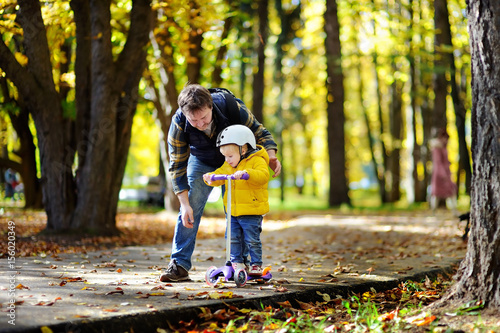 Middle age father showing his toddler son how to ride a scooter in a autumn park