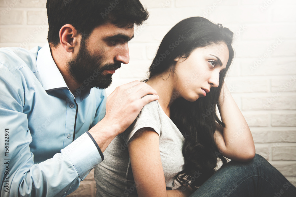 young woman looking away while man sitting behind her on the couch and consoling her young woman looking away while man sitting behind her on the couch and consoling her