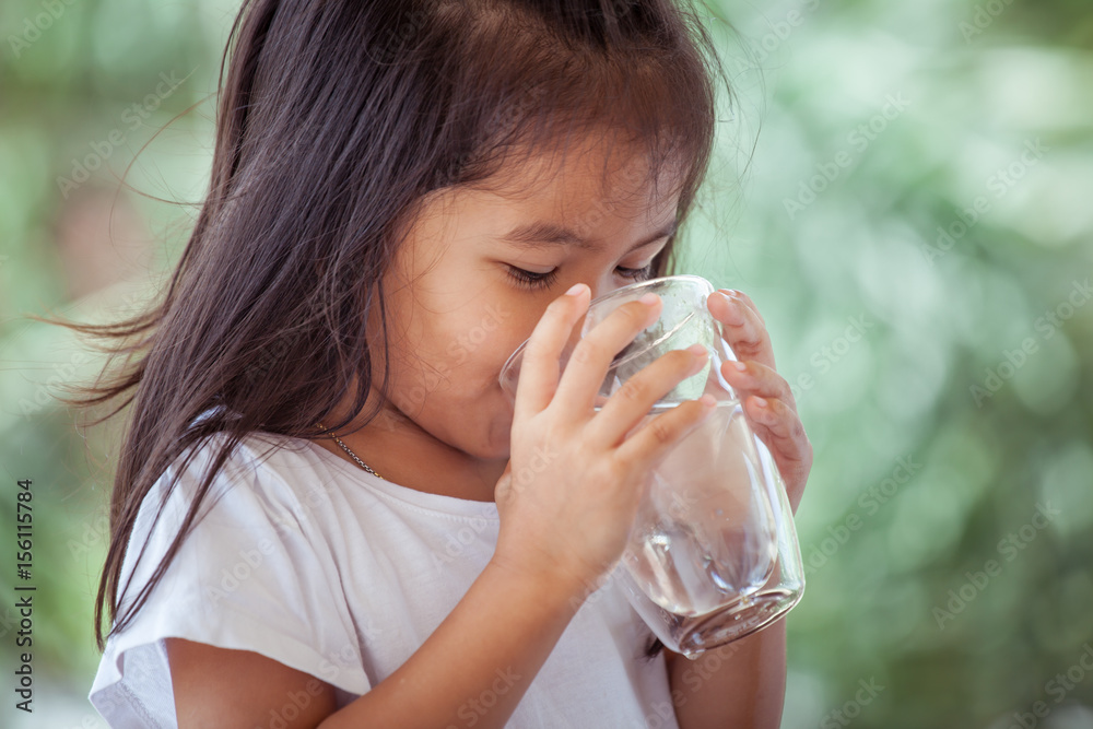 Cute asian little girl drinking fresh water from glass on green nature background Cute asian little girl drinking fresh water from glass on green nature background