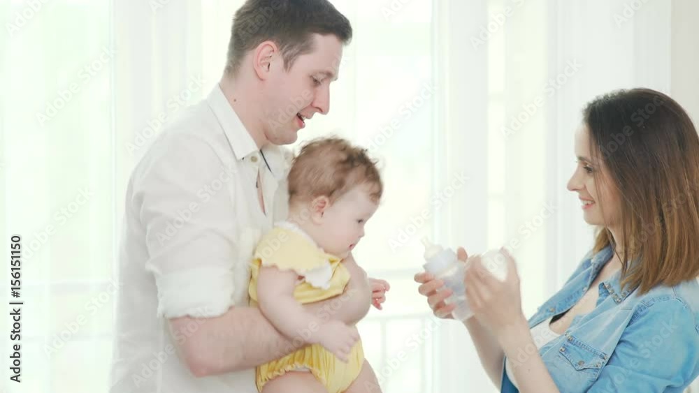 Young parents: father holds the baby in his arms, the mother feeds from the bottle. A young father holds his little daughter in his arms, a beautiful mother gives her daughter a drink from a bottle Young parents: father holds the baby in his arms, the mother feeds from the bottle. A young father holds his little daughter in his arms, a beautiful mother gives her daughter a drink from a bottle
