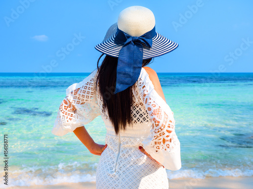 Woman portrait in white dress and sunhat standing on tropical beach, her arms raised and looking at sea on summer day.