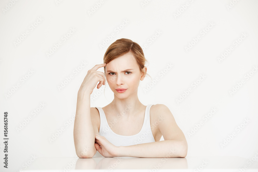 Young beautiful girl looking at camera thinking sitting at table over white background. Facial treatment. Young beautiful girl looking at camera thinking sitting at table over white background. Facial treatment.
