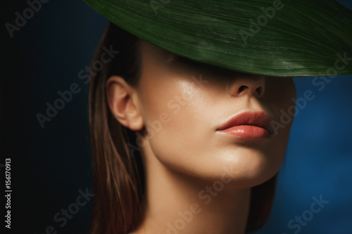Closeup of young woman covering face behind fresh green leaf.