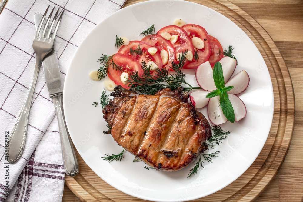 Pork steak cooked on the grill is decorated with a tomato, radish, dill, parsley, greens of garlic, mint with a fork and knife on a checkered napkin