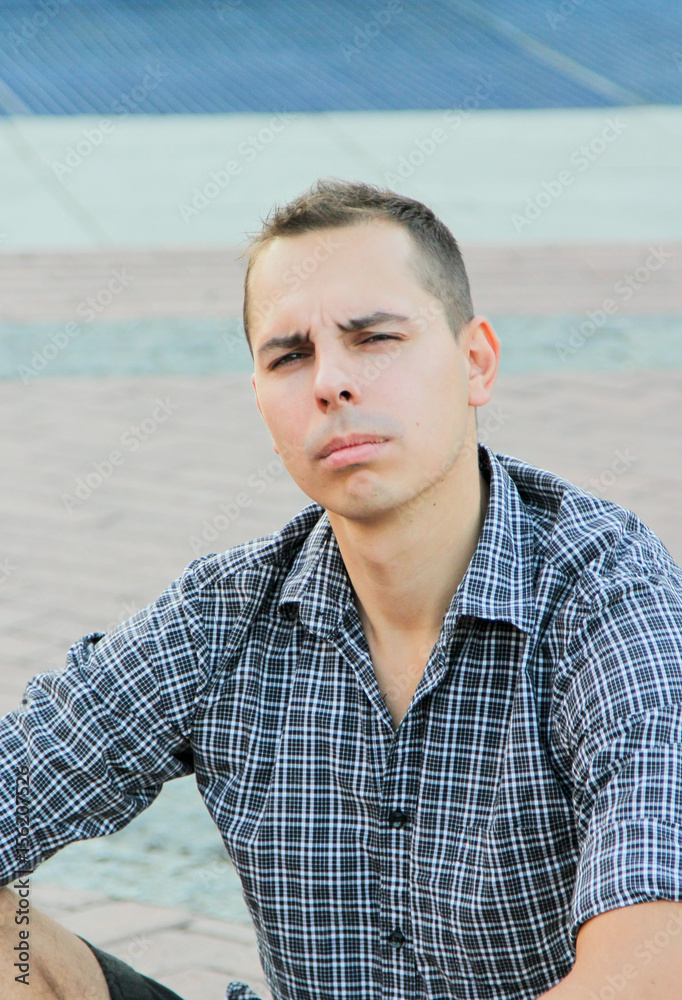 Man in shirt sitting on the pavement. Human emotions. Frown young man looking at the camera Man in shirt sitting on the pavement. Human emotions. Frown young man looking at the camera