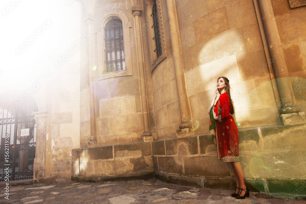 Stunning woman in red sari poses before stone wall in the morning lights