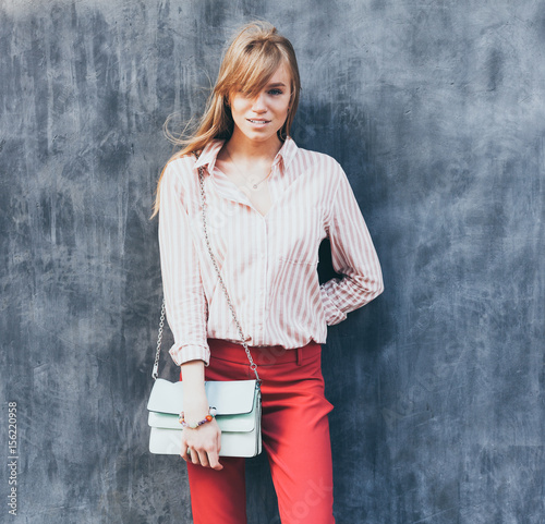 Portrait of a young woman dressed in a blouse, red Chino Trouser, a handbag turquoise on her shoulder. Posing next to a gray wall. photo