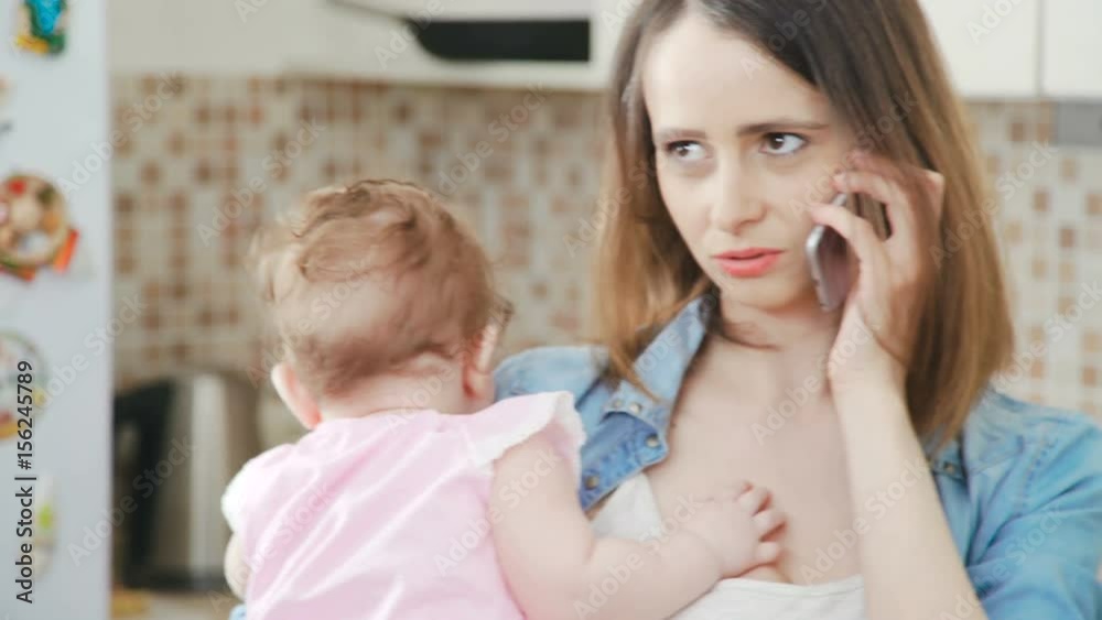 A beautiful and young mother is holding her little baby in her arms while talking on the phone. Modern mom holds a daughter in her arms and uses a mobile phone. Young woman with baby and phone. A beautiful and young mother is holding her little baby in her arms while talking on the phone. Modern mom holds a daughter in her arms and uses a mobile phone. Young woman with baby and phone.