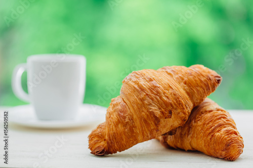 Tasty buttery croissants and cup of coffee on white wooden table over green tree background. Traditional french breakfast