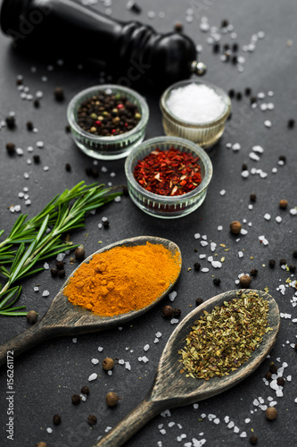 Close-up On a stone black spice surface, on a wooden spoon of turmeric, in glass pialls salt, paprika, dried basil, pepper mill, bottle with olive oil, rosemary, colorful, top view