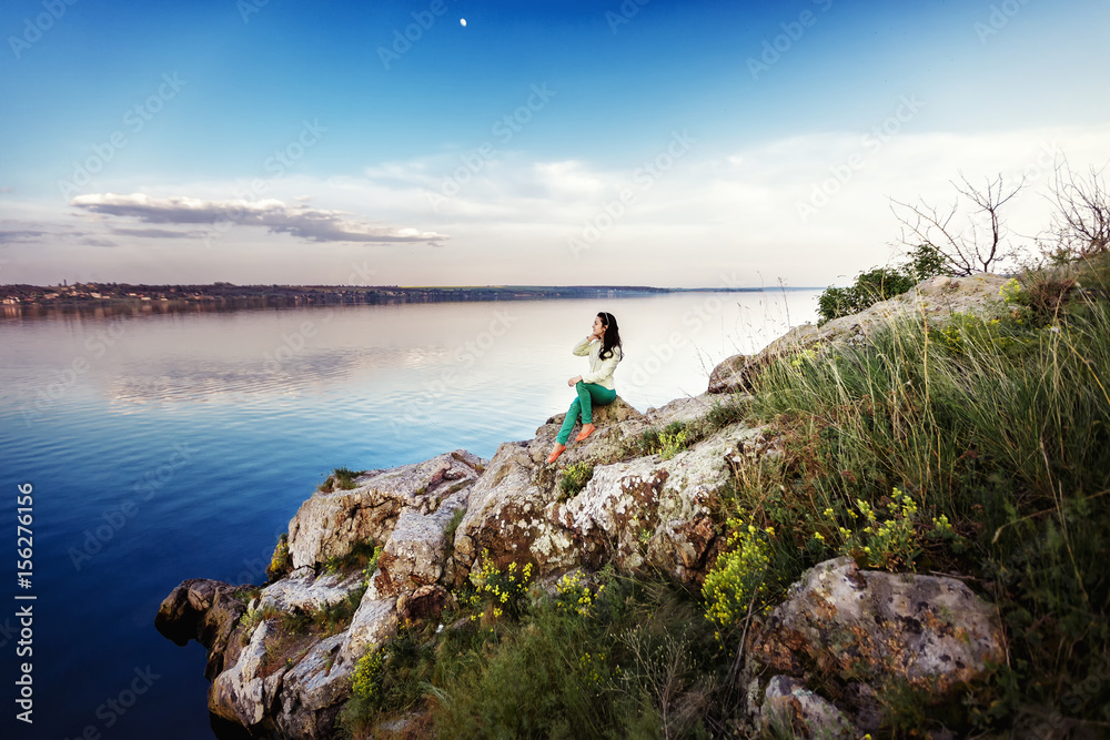 Young woman sits on rock and enjoys view of river at sunset Young woman sits on rock and enjoys view of river at sunset