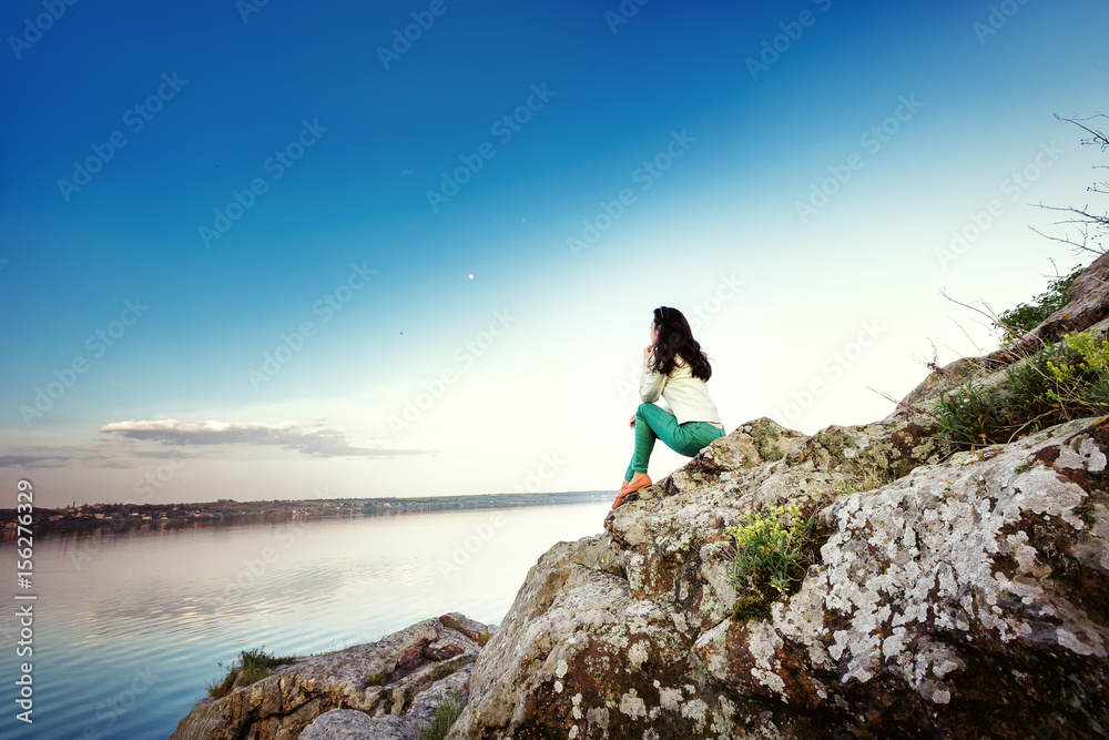 Young woman sits on rock and enjoys view of  river at sunset