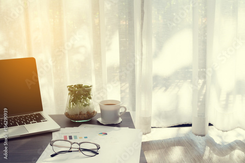 office table with blank screen on laptop, coffee cup, garden plant  pot and statistic report paper ob blurry drape  at living room, concept of business lifestyle.