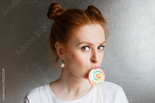 Redhead beautiful girl with multi-colored round candy and funny hairstyle on silver background.