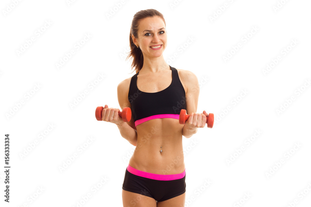 young smiling female athlete stands in front of the camera at the top and shorts and holding a dumbbell young smiling female athlete stands in front of the camera at the top and shorts and holding a dumbbell