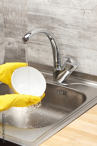 Hands in yellow gloves washing white bowl under tap water