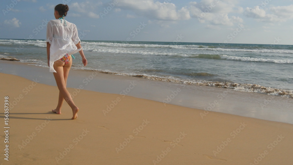 Beautiful woman in swimsuit and shirt walking on sea beach barefoot. Young girl going on the ocean shore. Female foot stepping on the sand with sea waves background. Summer vacation concept Rear view