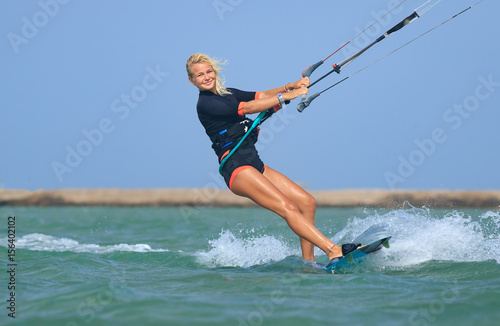 Kite surfing girl in sexy swimsuit with kite in sky on kiteboard in the blue sea riding waves saying hi. Recreational activity, water sports, action, hobby and fun in summer time. Kiteboarding sport