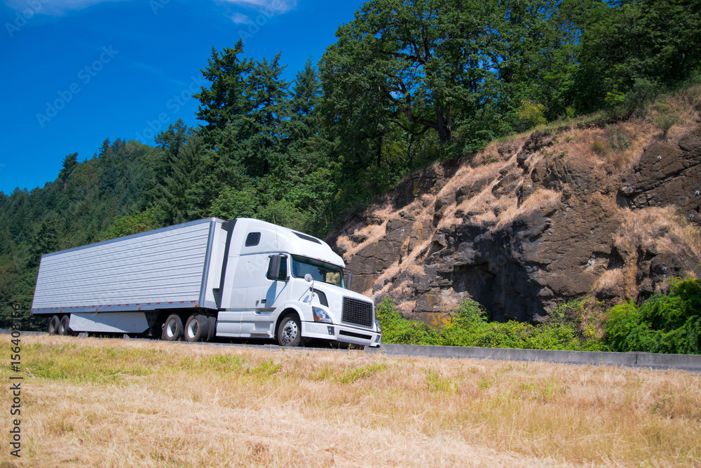 White semi truck trailer going highway with rocks green trees White semi truck trailer going highway with rocks green trees