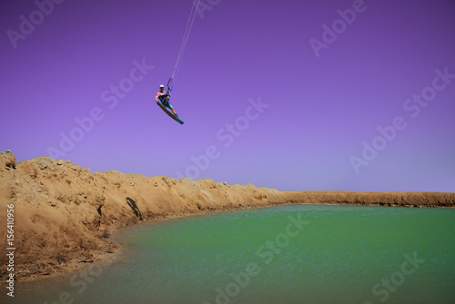 Professional kitesurfer sportsman with kite in sky jumps high acrobatics kiteboarding trick with grab of kiteboard to the green water lagoon. Recreational activity, extreme active air sports