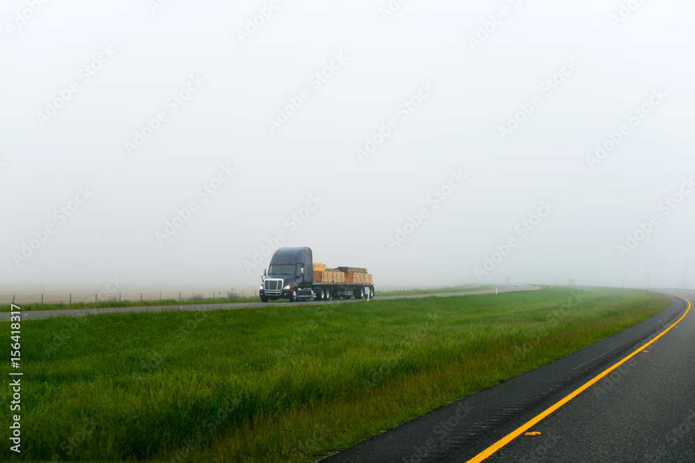 Dark big rig semi truck flat bed trailer lumber cargo foggy road with green grass