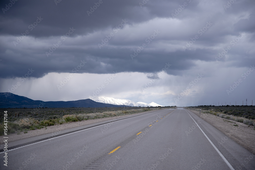 Naklejka premium Road stretches into horizon with stormy sky and snow-capped mountains