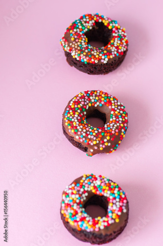 Three baked chocolate doughnuts with chocolate glaze with confetti over pink background. Overhead view.