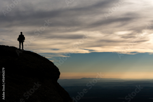 Black silhouette view of hiker man standing on high cliff watching ambient panorama. Cloudy sky and bright sunset on background.