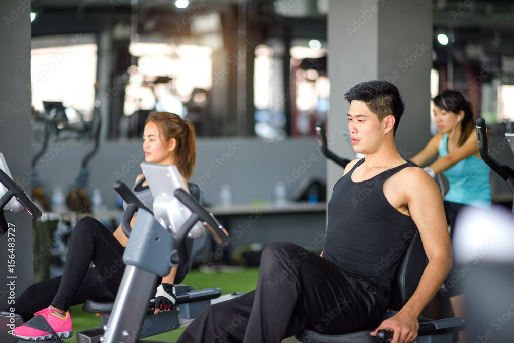 man riding stationary bicycles in modern gymnasium with woman doing the same action in background