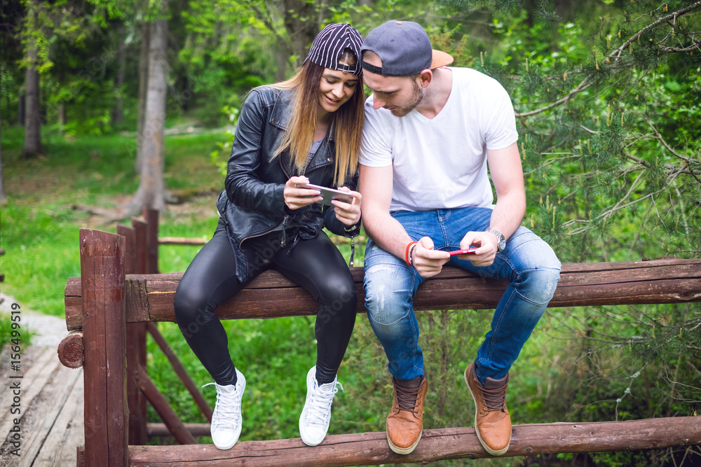 Young, modern couple holding a mobile phones and laughing. Concept of modern relationships. Close up of hipster people sitting on a wood in forest.