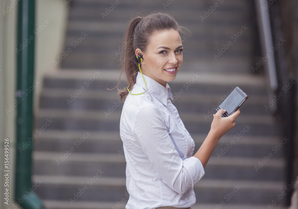 Youth Concepts and Ideas. Stylish Modern Caucasian Brunette Woman Posing Outdoors With Smartphone Gadget and Wearing Wireless Headphones. Youth Concepts and Ideas. Stylish Modern Caucasian Brunette Woman Posing Outdoors With Smartphone Gadget and Wearing Wireless Headphones.
