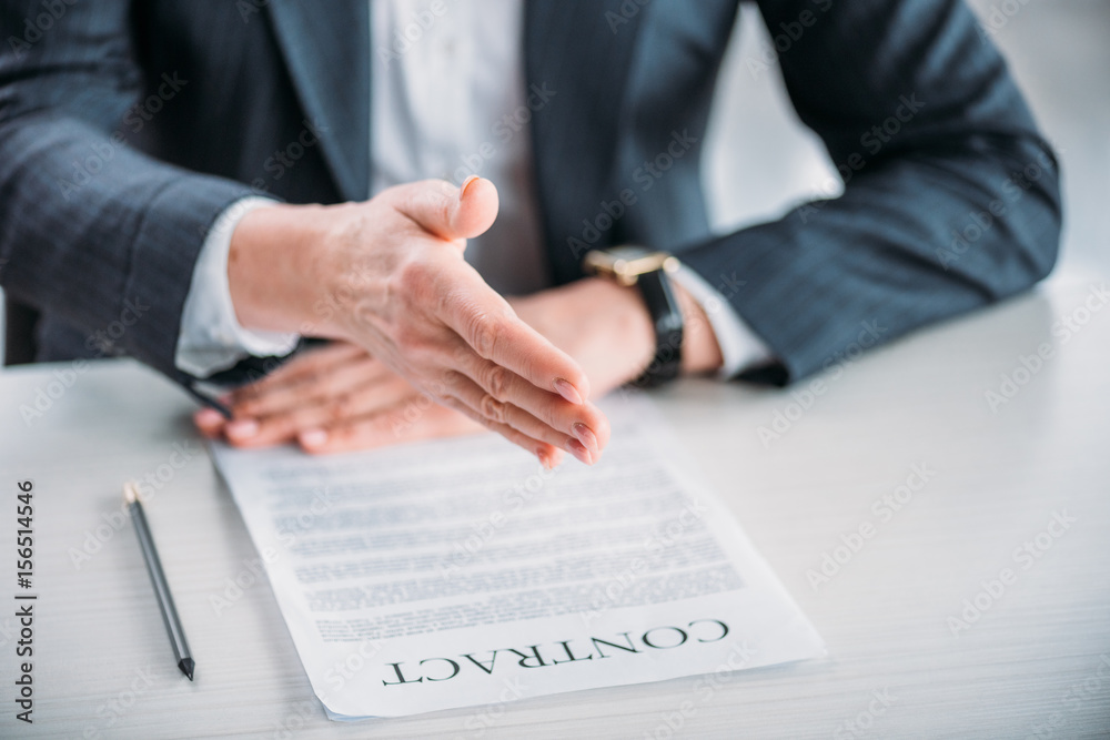 Partial view of businesswoman extending hand for handshake with contract on tabletop Partial view of businesswoman extending hand for handshake with contract on tabletop