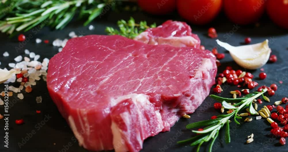 Beautiful juicy fresh meat steak on a table with salt, rosemary, garlic, and tomato on a black background, top view. Concept: fresh & natural products, bio products, meat products, organic, nutrition. Beautiful juicy fresh meat steak on a table with salt, rosemary, garlic, and tomato on a black background, top view. Concept: fresh & natural products, bio products, meat products, organic, nutrition.