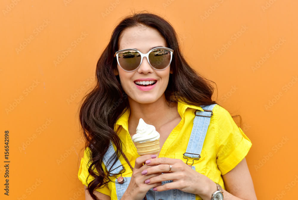 Beautiful  funny young hipster teen girl eating ice cream cone. laughs happy. Bright casual wear, denim shorts. Orange  background, urban style, sunglasses